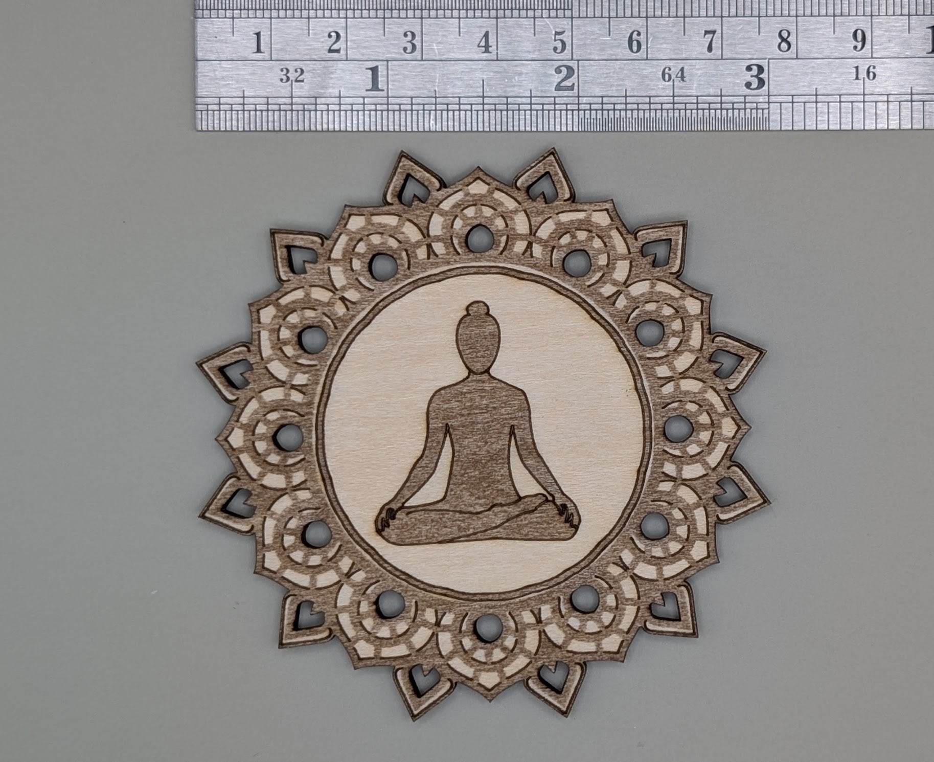 Decorative wooden ornament with meditating figure and mandala design, measured against a ruler.