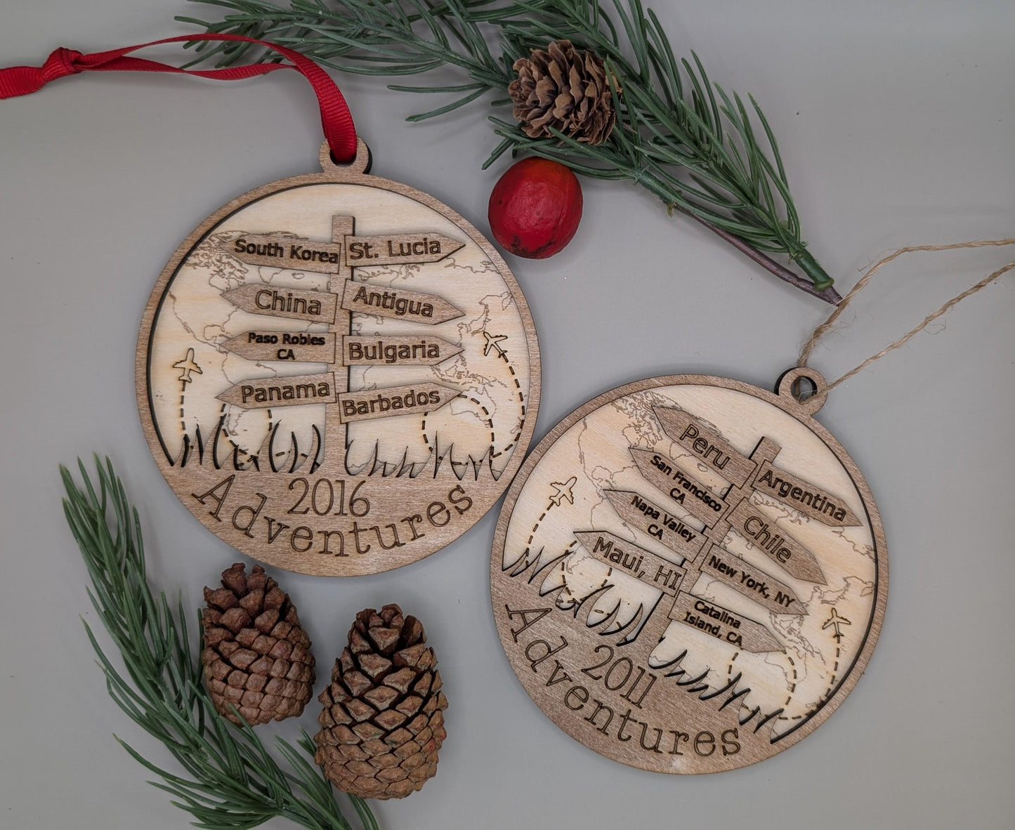 Two wooden Christmas ornaments with engraved travel destinations and dates, surrounded by pine cones and a red ribbon.