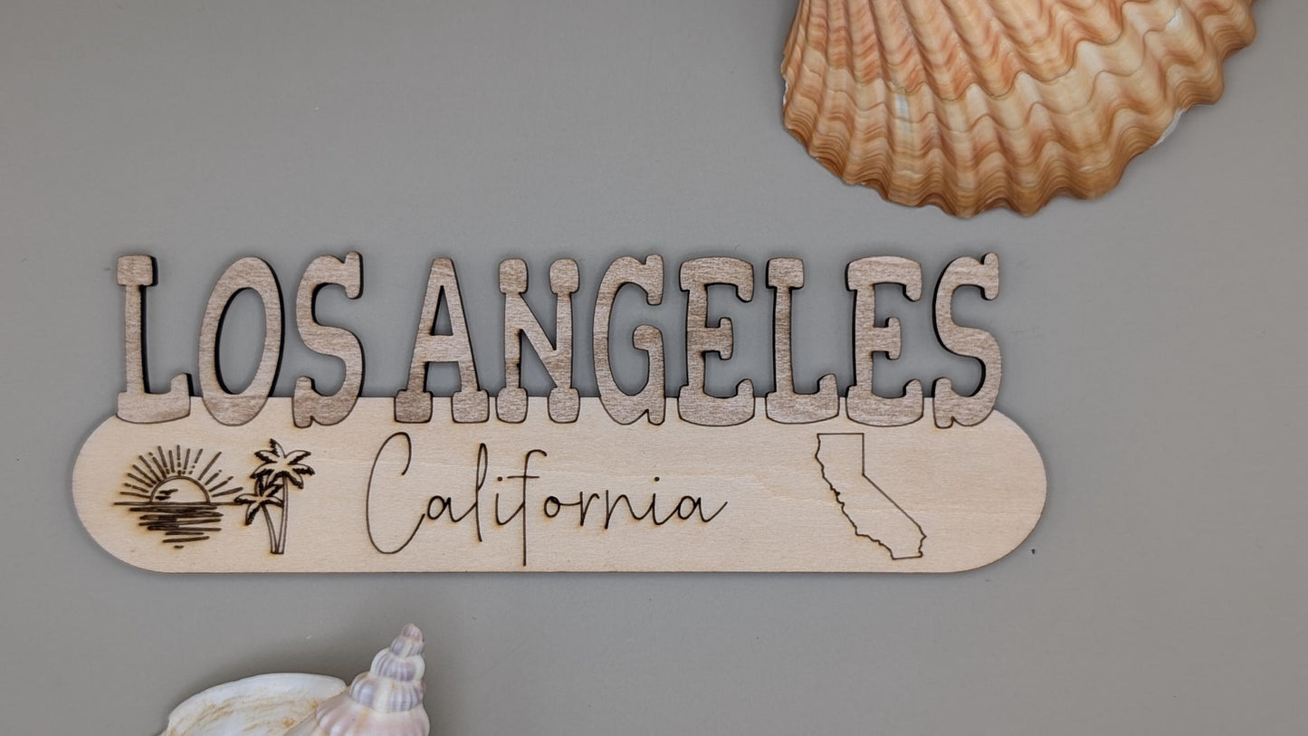 Wooden sign with 'Los Angeles California' on a gray background with shells.