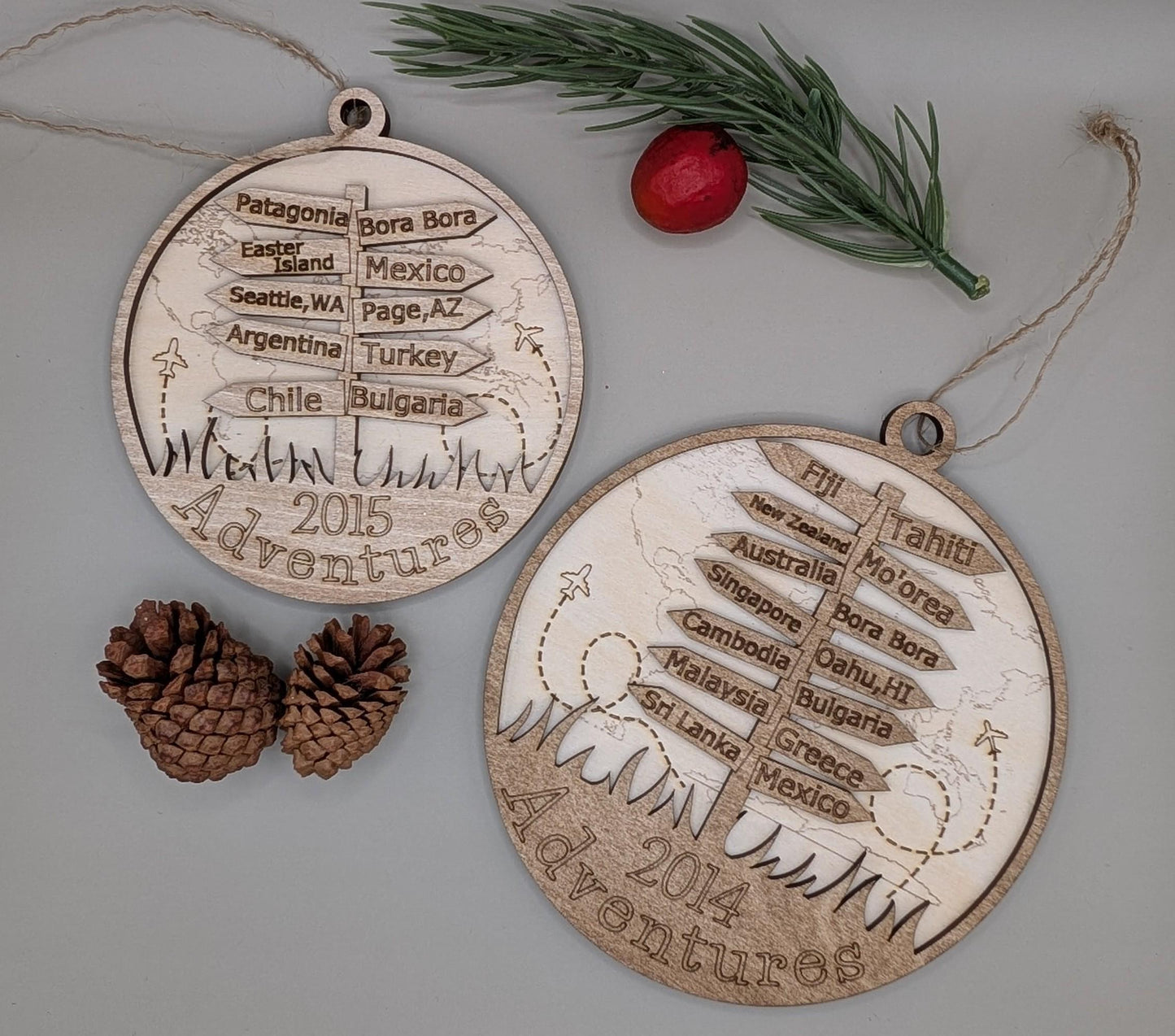 Two wooden Christmas ornaments with engraved travel destinations on a light background with pine cones and a red berry.