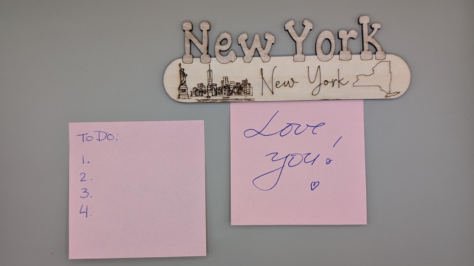 Decorative sign with 'New York' and a silhouette of the Statue of Liberty, two pink sticky notes on a gray wall.